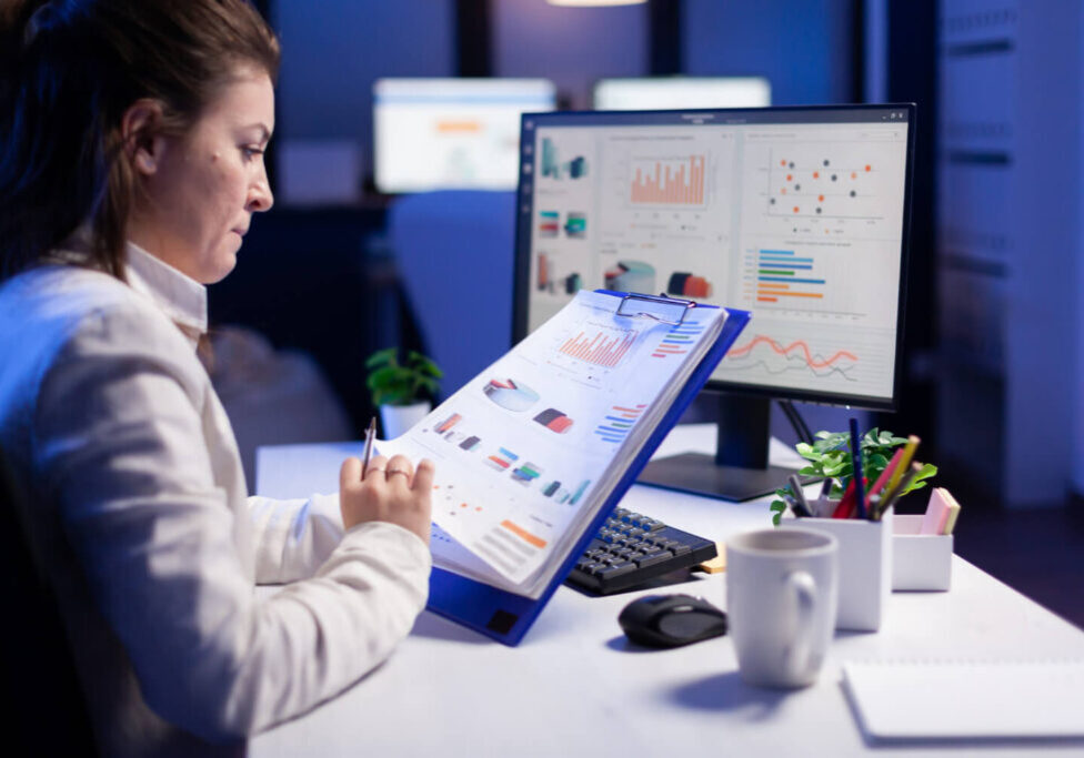 Freelancer woman comparing graphics from clipboard with grafics from computer at business office